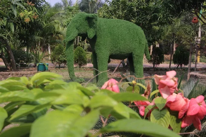 A life-size green topiary elephant among palms, with pink flowers in front